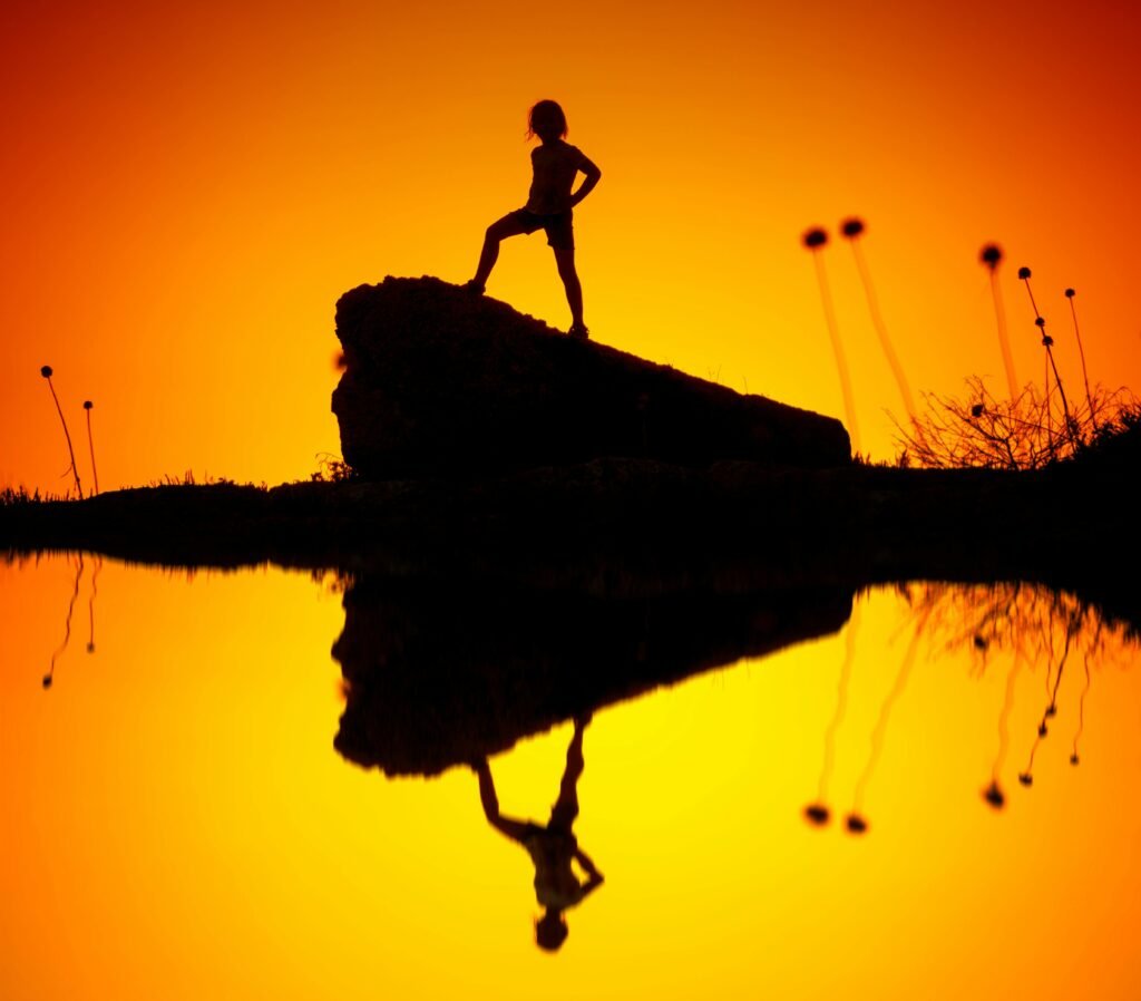 A child's silhouette on a rock with reflection in water during a vibrant sunset.